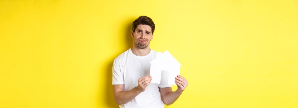 Real estate concept. Displeased young man showing paper house model and grimacing upset, standing over yellow background.