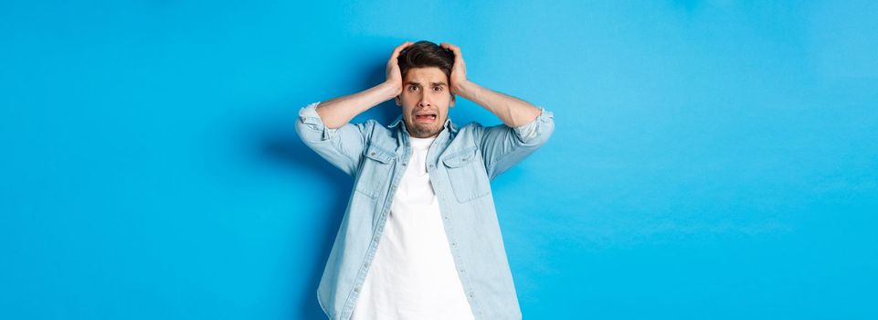 Image of man in panic holding hands on head, looking frustrated and anxious, standing against blue background.