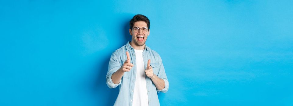 Confident man saying congrats, winking and pointing at you, standing pleased over blue background and smiling.