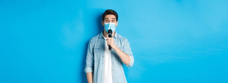 Young man in medical mask giving speech, holding microphone and looking confused, standing over blue background.