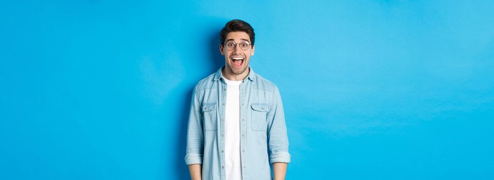 Attractive happy man in glasses looking surprised, checking out advertisement, standing over blue background.