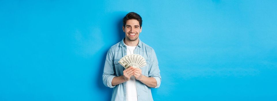 Handsome smiling man holding money, concept of finance and banking, standing over blue background.