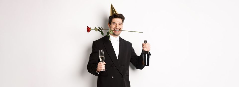 Funny guy in trendy suit, celebrating and having a party, holding rose in teeth and champagne, standing over white background.