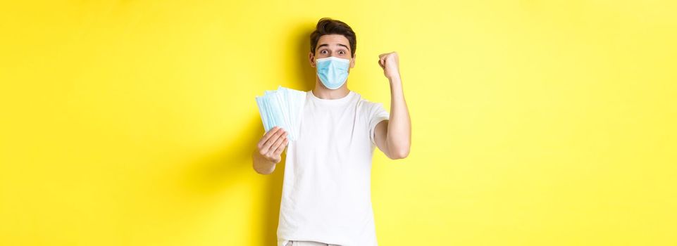 Concept of covid-19, quarantine and preventive measures. Happy man triumphing, raising hand to celebrate something and giving medical mask, standing against yellow background.