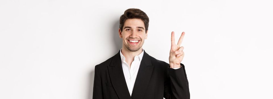 Close-up of handsome businessman in black suit, smiling amazed, showing number two, standing over white background.