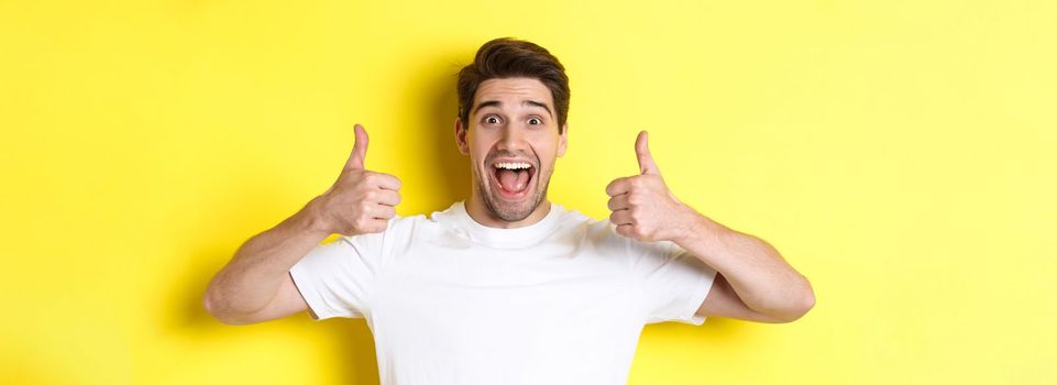 Close-up of handsome young man showing thumbs up, approve and agree, smiling satisfied, standing over yellow background.