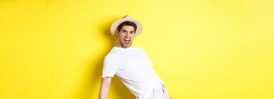 Concept of tourism and vacation. Excited young man tourist celebrating, shouting for joy and dancing, standing over yellow background.