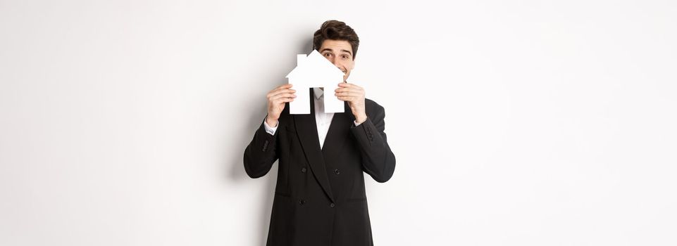 Image of handsome broker in black suit, showing house maket and smiling, selling homes, standing against white background.