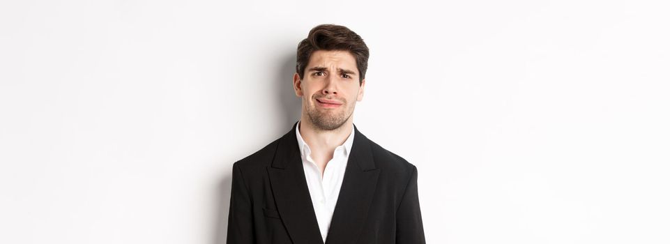 Close-up of confused handsome man in suit, grimacing and looking perplexed, wearing trendy suit, standing against white background.