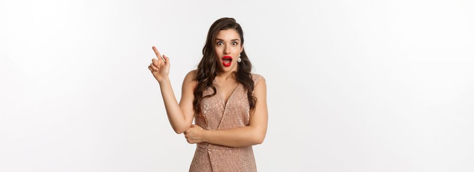 Surprised beautiful woman in luxurious dress and red lips, pointing finger right at promo and starring amazed at camera, standing over white background.