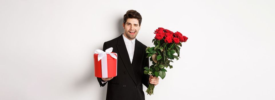 Concept of holidays, relationship and celebration. Handsome and confident man in black suit, going on a date, holding bouquet of roses and present, standing against white background.