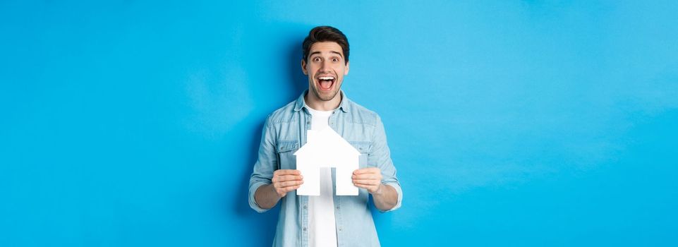 Insurance, mortgage and real estate concept. Happy man holding house model and smiling excited, buying property or renting apartment, standing against blue background.