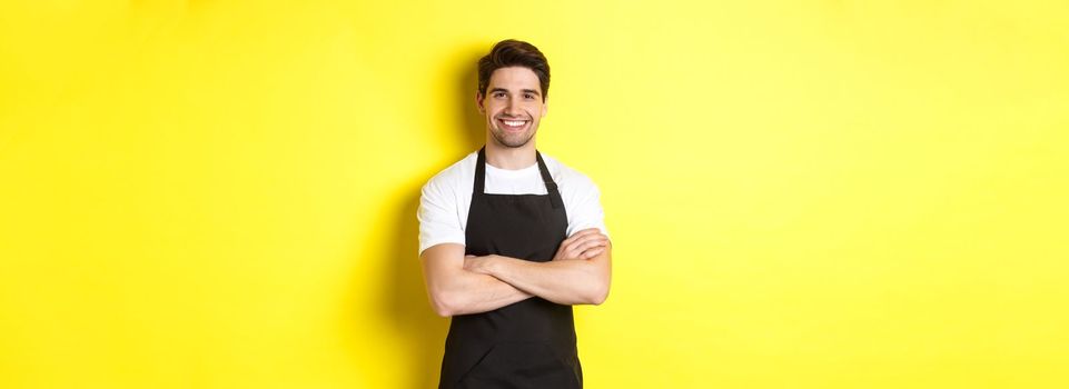 Smiling male waiter in black apron standing confident, cross arms on chest against yellow background.