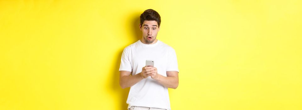 Man looking surprised in smartphone, reading message on cell phone, standing in white outfit against yellow background. Copy space