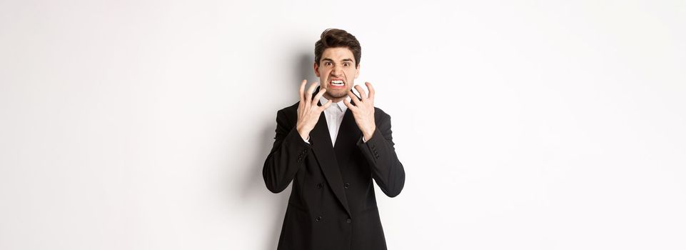 Image of angry businessman in suit, looking with furious expression and clenching fists, express hatred, standing mad over white background.