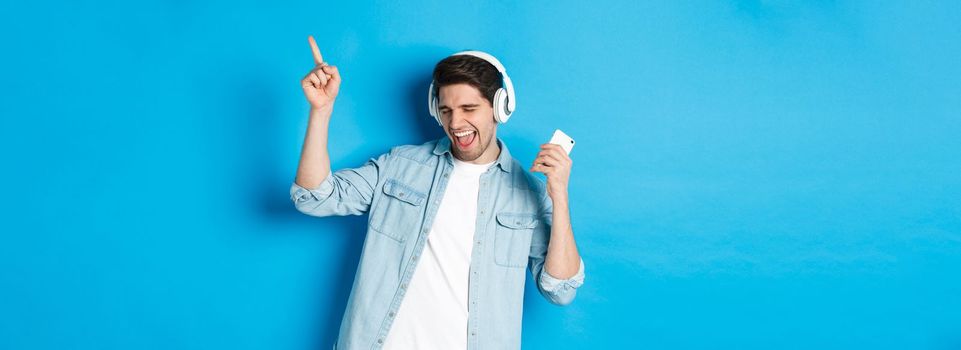 Happy handsome man dancing, listening music on smartphone in headphones, standing over blue background.