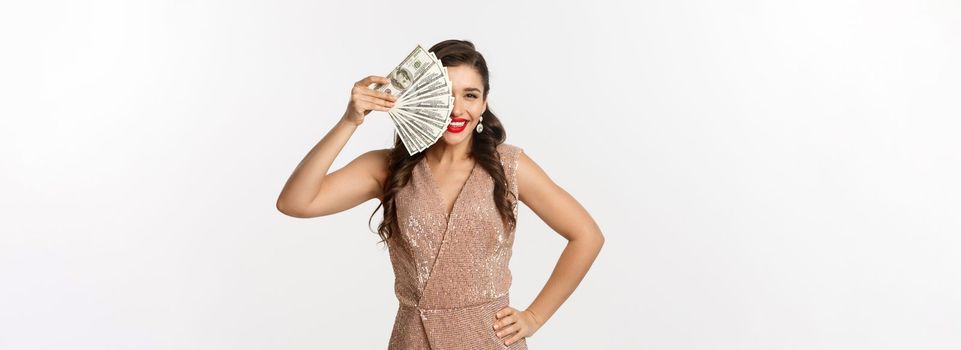 Shopping concept. Beautiful and happy woman in elegant dress, holding money on face and smiling pleased, standing over white background.
