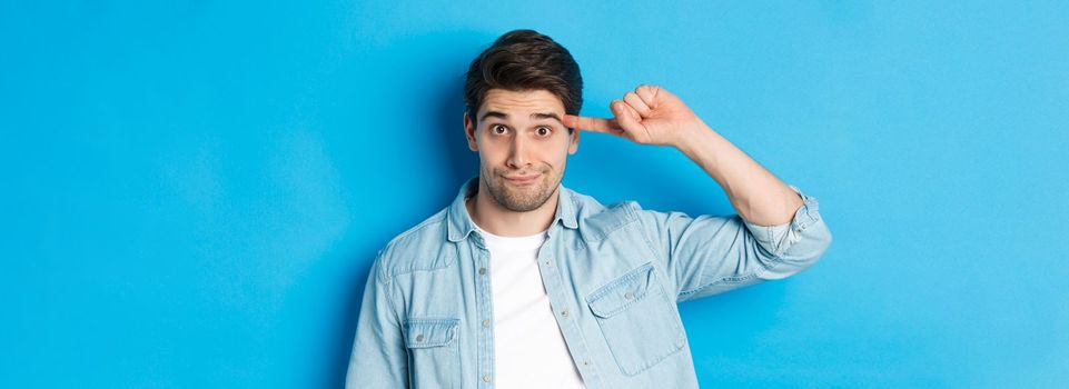 Close-up of man scolding for acting stupid or crazy, rolling finger on head and looking at camera, standing over blue background.