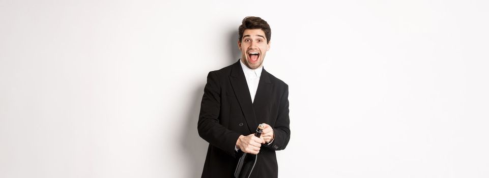 Image of attractive man in black suit having a party, celebrating new year and opening bottle of champagne, standing happy against white background.