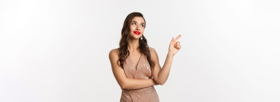 Christmas, holidays and celebration concept. Dreamy beautiful woman in party dress, pointing and looking left, thinking about new year party, white background.