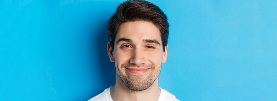 Headshot of attractive man smiling pleased, looking intrigued, standing over blue background.