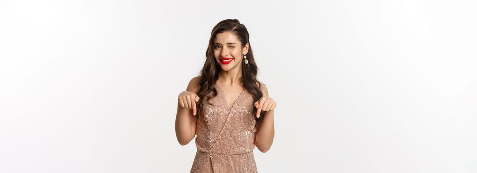 Elegant female model with red lips and earrings, pointing fingers down at christmas offer, winking and smiling, showing promo, standing in party dress over white background.