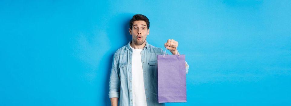 Concept of shopping, holidays and lifestyle. Handsome surprised guy holding paper bag from shop and looking amazed, standing over blue background.