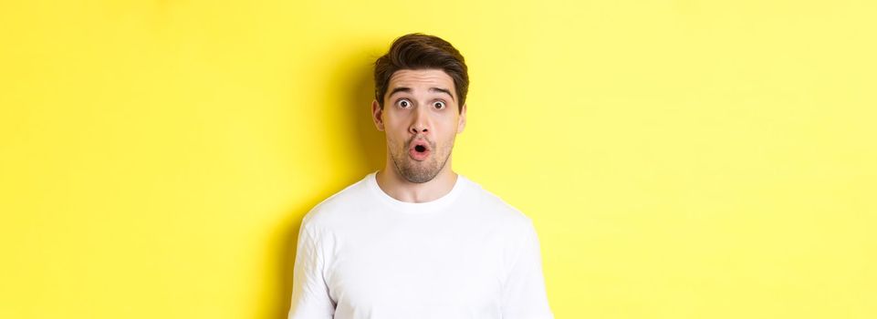 Close-up of handsome caucasian man saying wow, looking impressed, standing against yellow background.