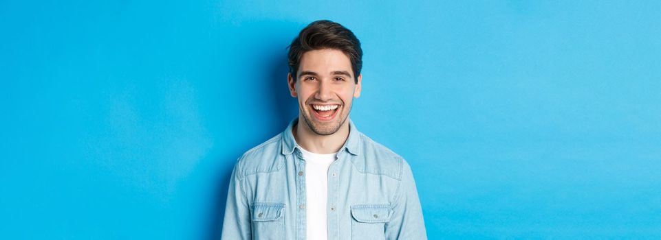 Close-up of handsome young man laughing, wearing casual clothes, standing over blue background.
