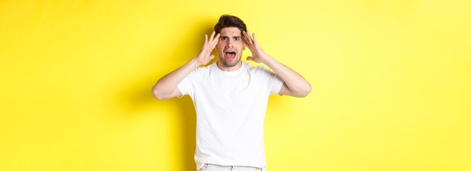 Frustrated guy looking alarmed, holding hands on head and feeling confused, panicking, standing over yellow background.