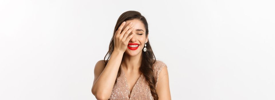Concept of New Year celebration and winter holidays. Fabulous woman with red lips, wearing party dress, laughing and looking delighted, standing over white background.