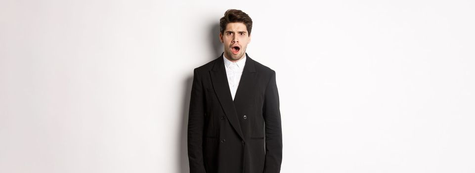 Portrait of shocked and startled handsome man in suit, drop jaw and looking in awe at camera, standing over white background.