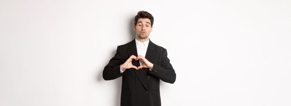 Image of handsome groom in black suit, showing heart sign, close eyes and pucker lips, waiting for kiss, standing against white background.