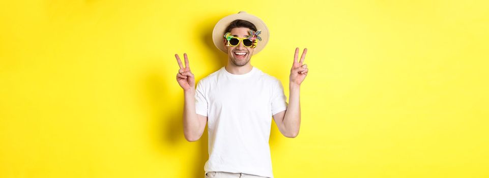 Concept of tourism and lifestyle. Happy man enjoying trip, wearing summer hat and sunglasses, posing with peace signs for photo, yellow background.