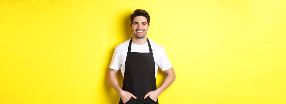 Happy barista in black apron looking at camera. Coffee shop owner wearing cafe uniform and smiling, standing over yellow background.