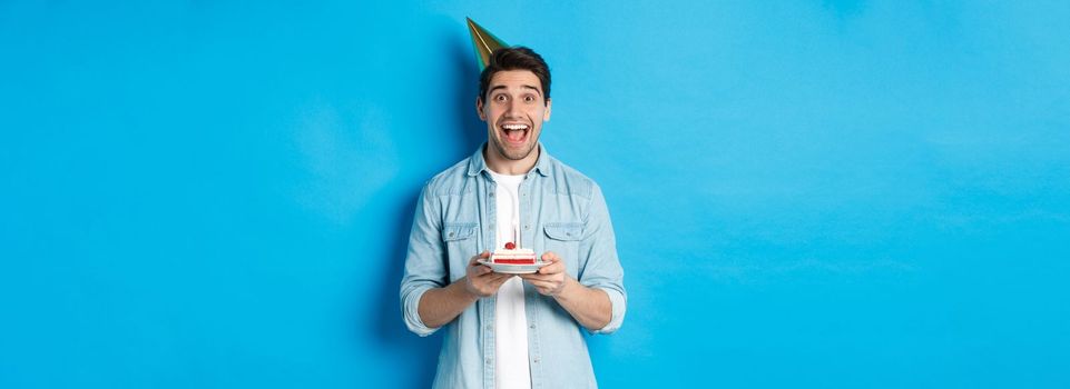 Cheerful young man celebrating birthday in party hat, holding b-day cake, standing against blue background.