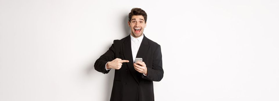 Image of cheerful businessman looking amazed, pointing at mobile phone, standing in suit over white background.