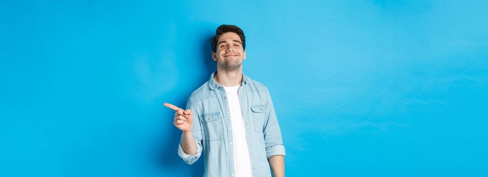 Confident handsome guy smiling sassy, pointing finger right at copy space, showing advertisement, standing over blue background.
