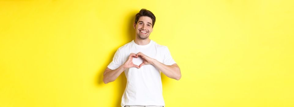 Happy romantic man showing heart sign, smiling and express love, standing over yellow background. Copy space