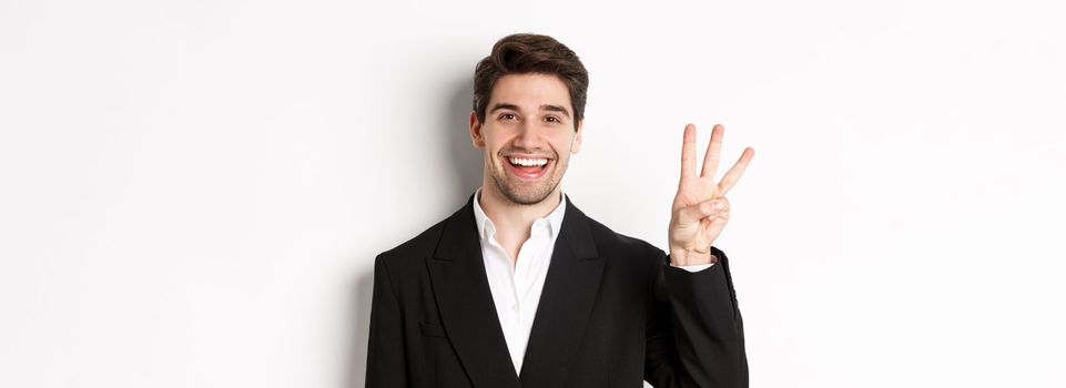 Close-up of handsome businessman in black suit, smiling amazed, showing number three, standing over white background.