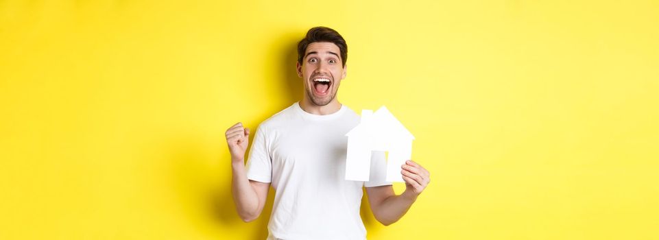 Real estate concept. Excited man holding paper house model and celebrating, standing happy over yellow background.