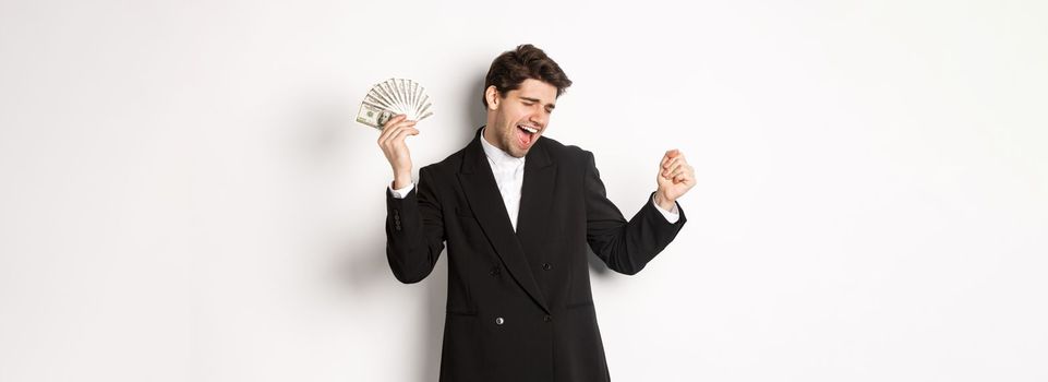 Portrait of handsome and successful businessman in suit, dancing with money, standing against white background.