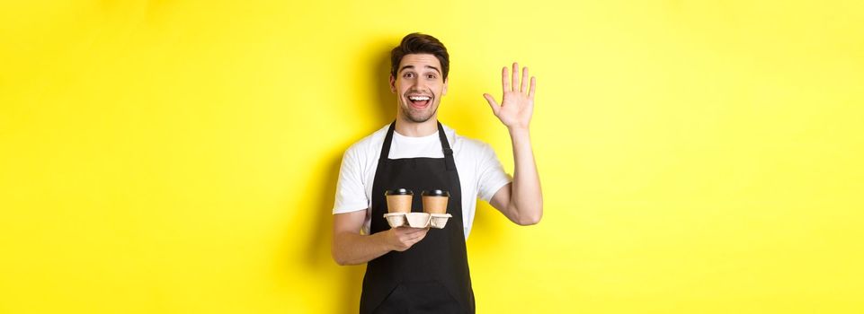 Friendly waiter in cafe waving hand at customer, holding takeaway coffee oder, standing against yellow background in black apron.