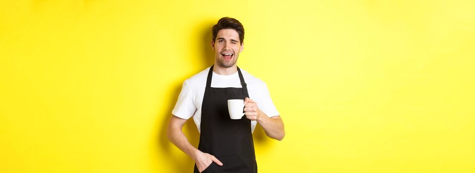 Cheerful barista in black apron serving coffee and winking, standing against yellow background.