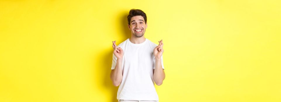 Hopeful smiling man holding fingers crossed, making wish or praying, standing over yellow background.