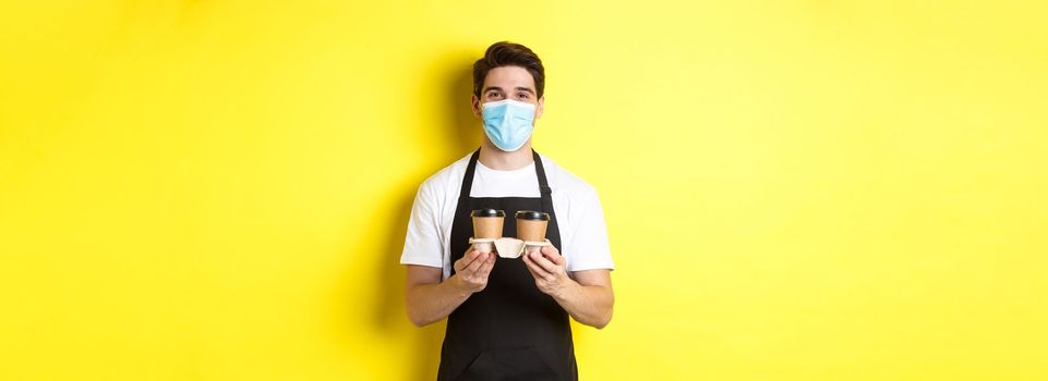 Concept of covid-19, cafe and social distancing. Barista in medical mask serving coffee in takeaway cups, standing in black apron against yellow background.