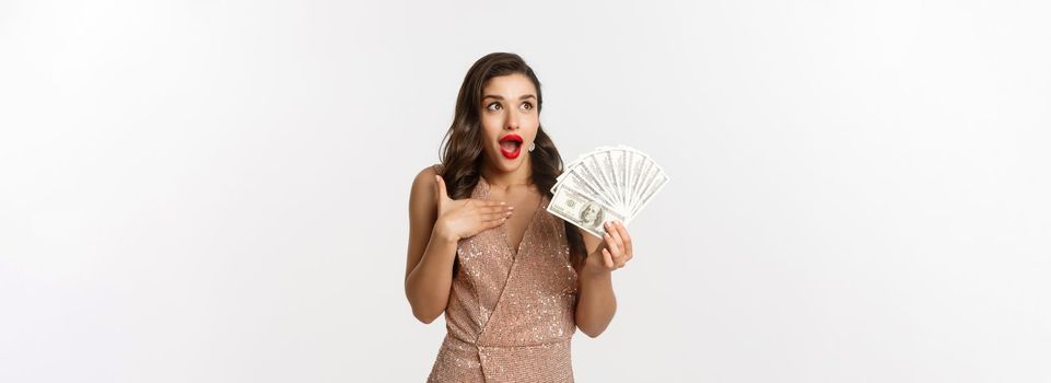 Shopping concept. Image of surprised and excited woman holding money, staring at upper left corner amazed, standing with dollars in glamour dress, white background.