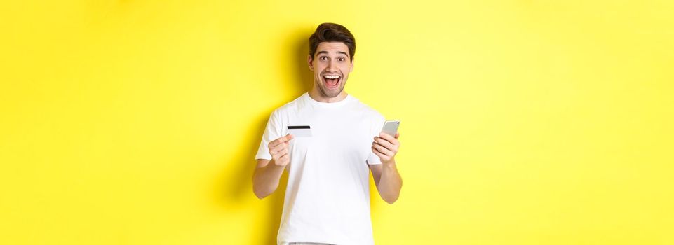 Happy male buyer holding smartphone and credit card, concept of online shopping in internet, standing over yellow background.