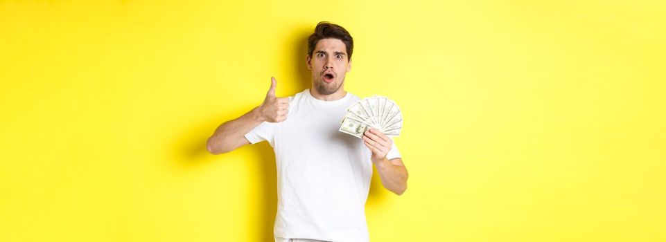 Impressed man showing thumb up, holding money credit, standing over yellow background.