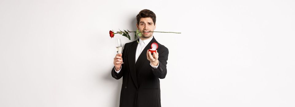 Romantic young man in suit making a proposal, holding rose in teeth and glass of champagne, showing engagement ring, asking to marry him, standing against white background.